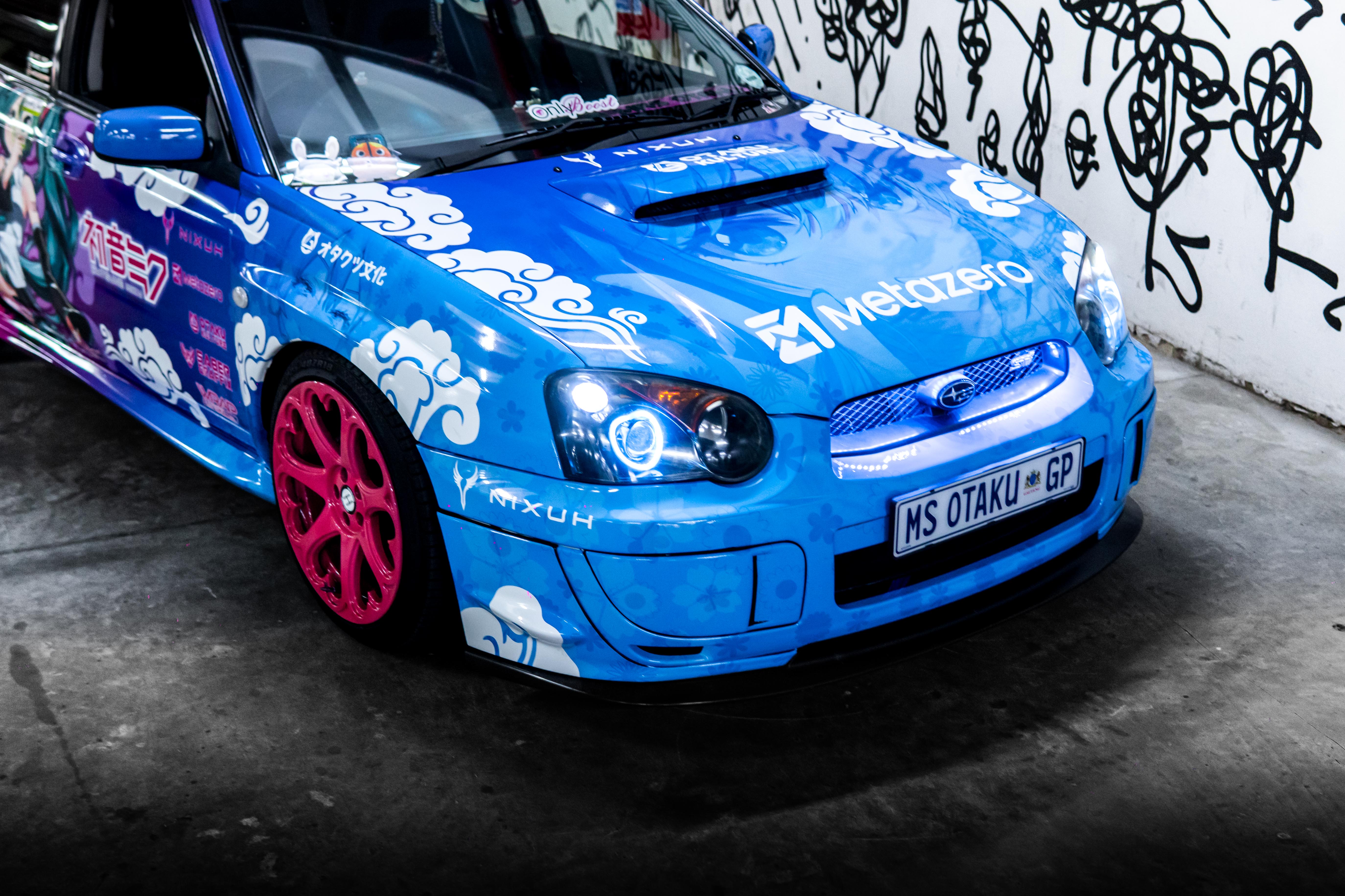 Blue/pink car with colorful graphics and branding in front of a graffiti-covered wall.