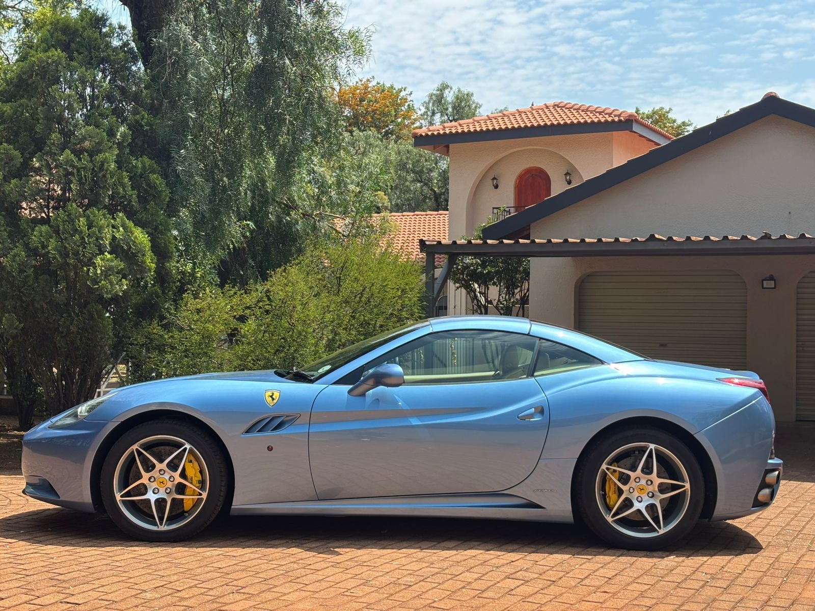 Blue Ferrari sports car parked on a driveway with a house and trees in the background