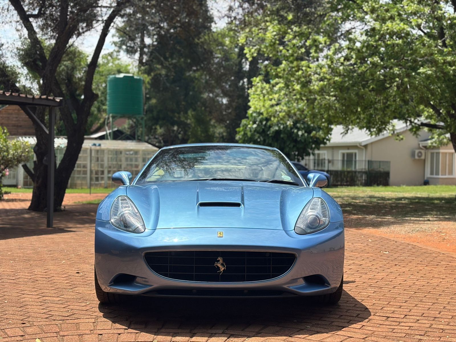 Blue Ferrari car parked on a brick driveway with trees and houses in the background
